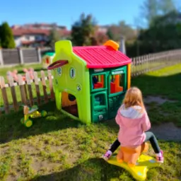 Dziewczynka siedzi na żółtym koniku na biegunach przed kolorowym, plastikowym domkiem z koszem do koszykówki na zewnątrz, na trawniku w słoneczny dzień.