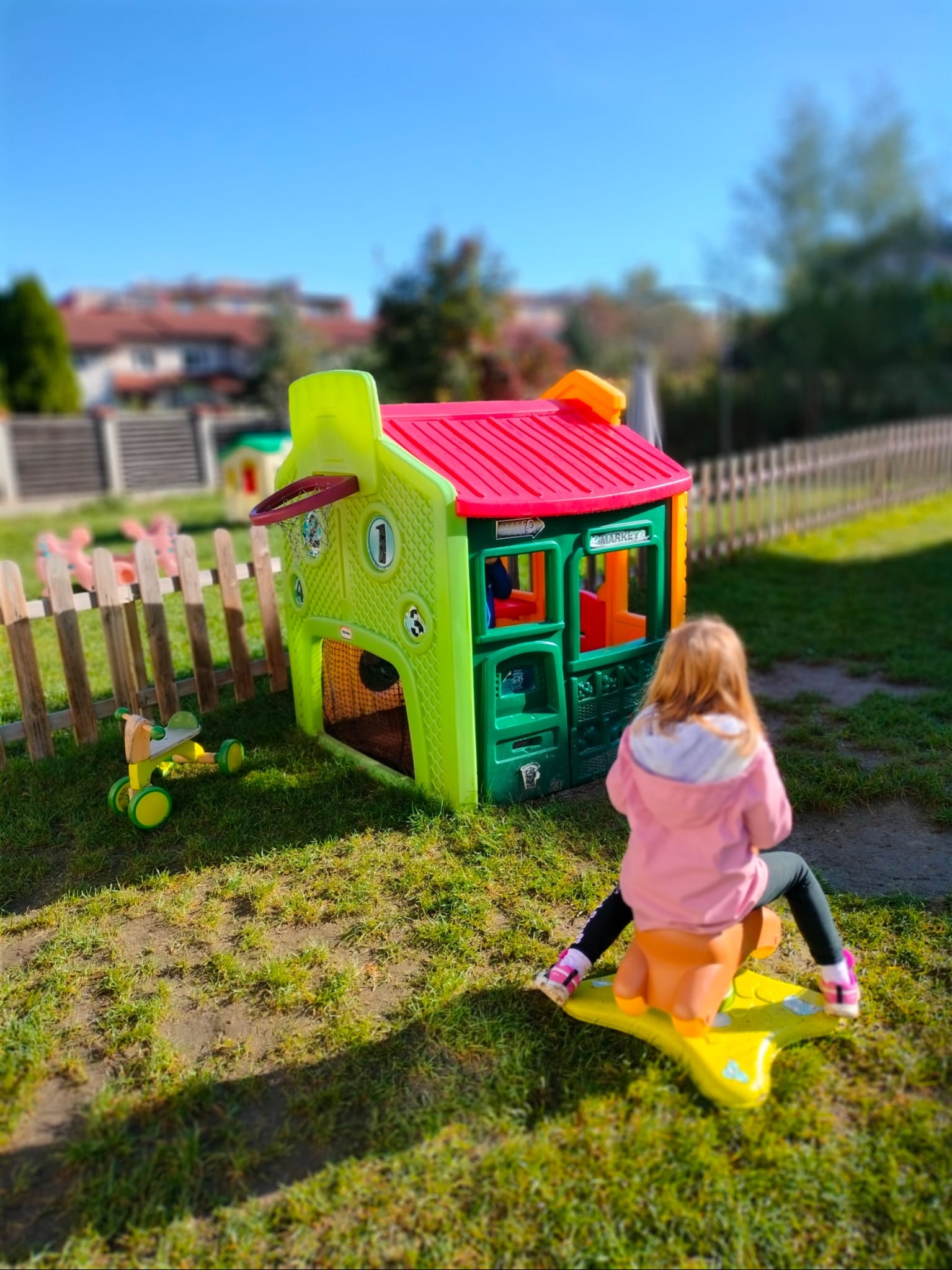 Dziewczynka siedzi na żółtym koniku na biegunach przed kolorowym, plastikowym domkiem z koszem do koszykówki na zewnątrz, na trawniku w słoneczny dzień.