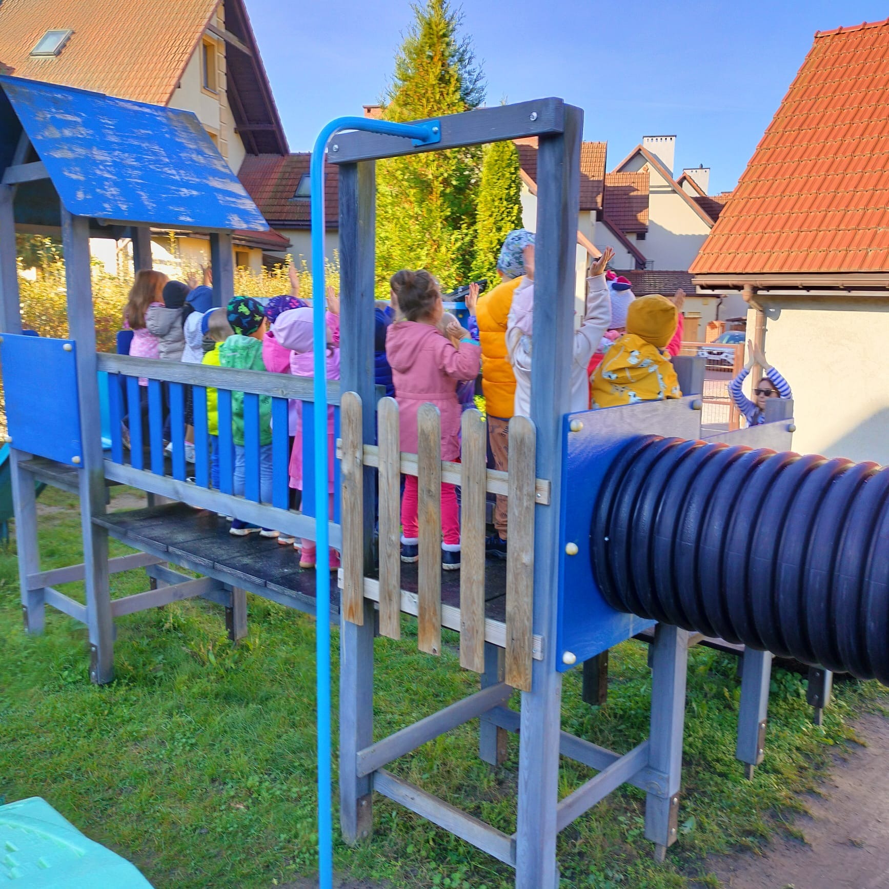 Grupa dzieci na drewnianym placu zabaw z tunelem, część dzieci macha rękami, w tle budynki mieszkalne z czerwonymi dachami.