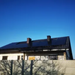 Dom jednorodzinny z zamontowanymi panelami słonecznymi na dachu, widoczny betonowy płot w cieniu drzewa i dwa kominy.