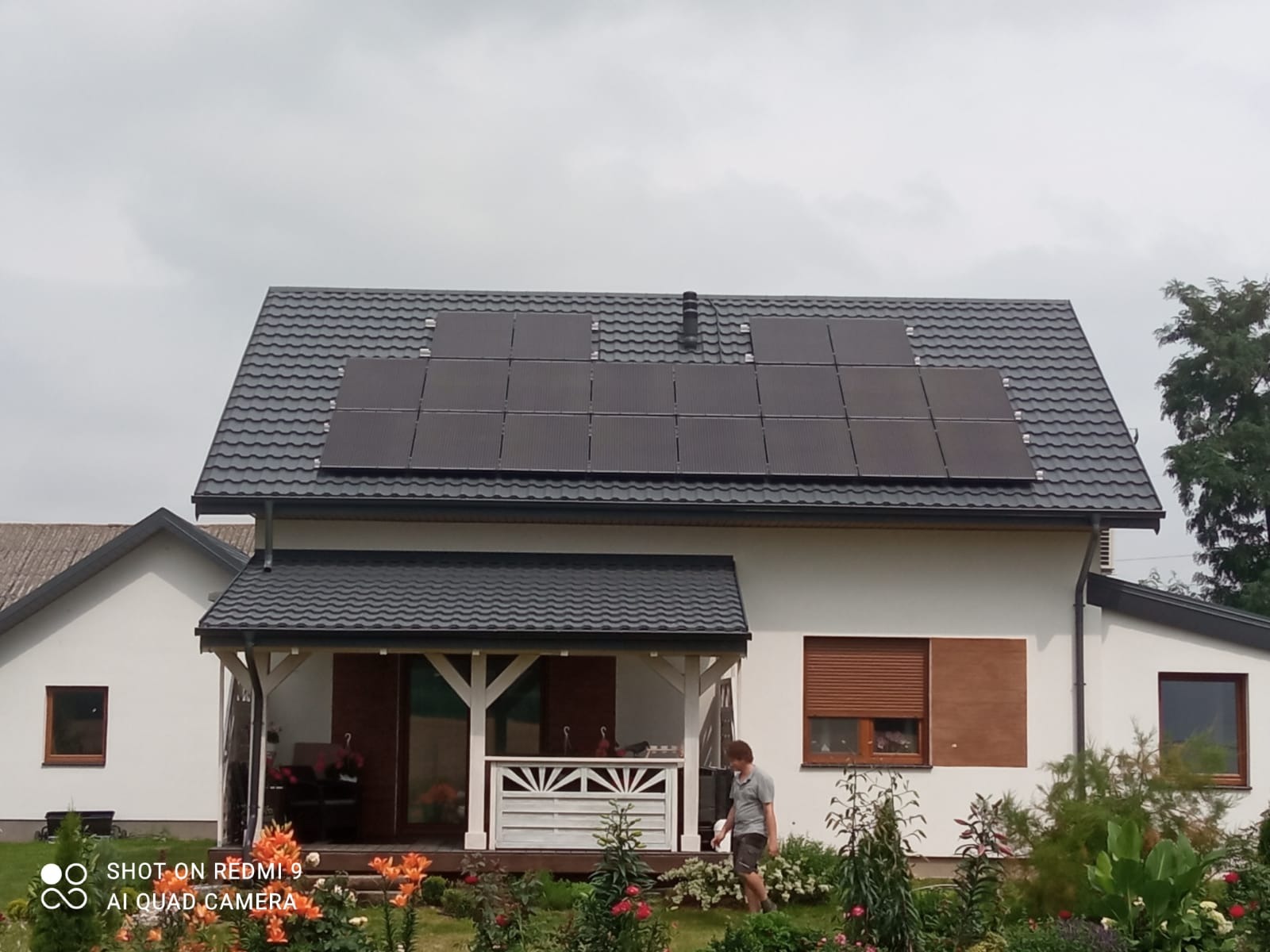 Dom jednorodzinny z zainstalowanymi panelami fotowoltaicznymi na dachu z blachodachówki, widok z ogrodu z kwitnącymi liliami, mężczyzna przechadza się w tle.