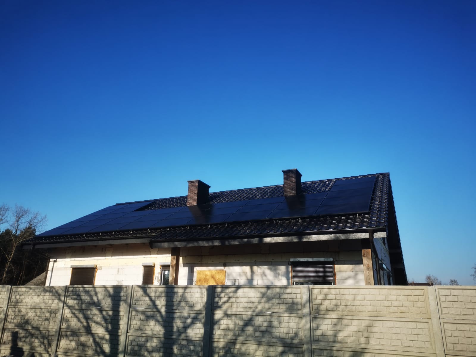 Dom jednorodzinny z zamontowanymi panelami słonecznymi na dachu, widoczny betonowy płot w cieniu drzewa i dwa kominy.