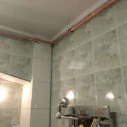 Miedziane rury instalacji wodnej poprowadzone pod sufitem w łazience z zielonymi płytkami, widoczne łączenia i uchwyty mocujące.