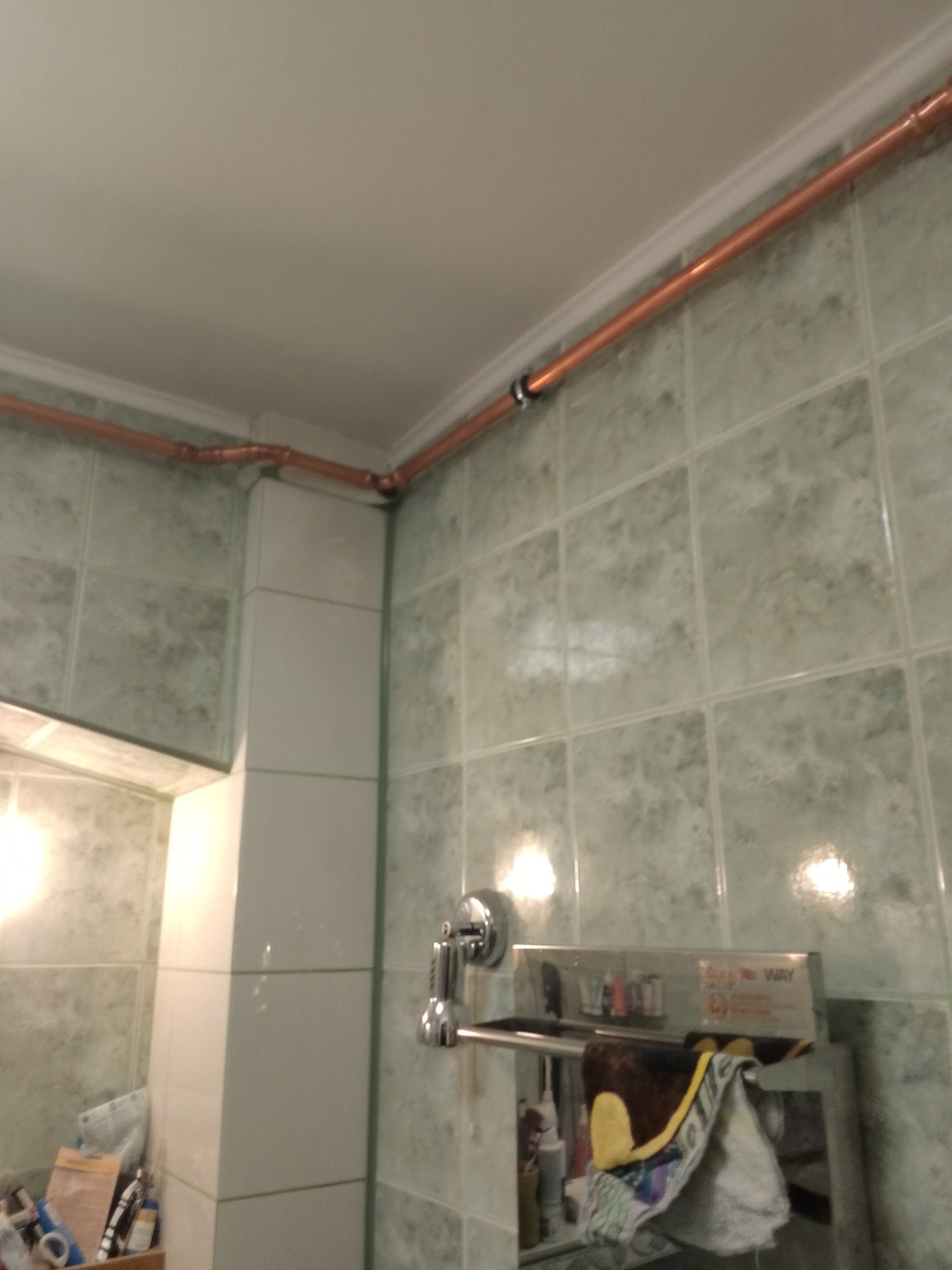 Miedziane rury instalacji wodnej poprowadzone pod sufitem w łazience z zielonymi płytkami, widoczne łączenia i uchwyty mocujące.