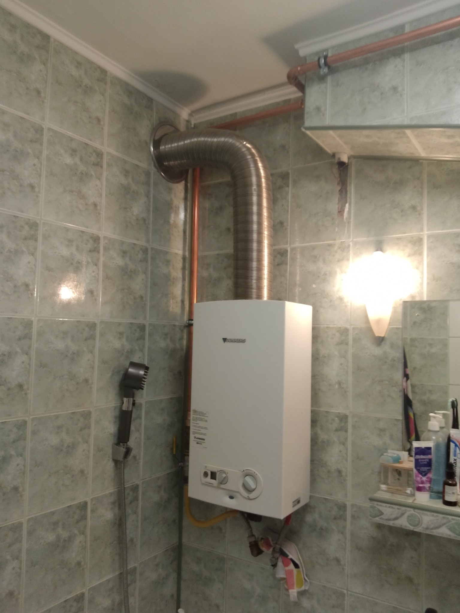 Biały piec gazowy Junkers z podłączonym, elastycznym kominem w łazience z zielonymi, kwadratowymi płytkami, widoczne rury miedziane i prysznic.