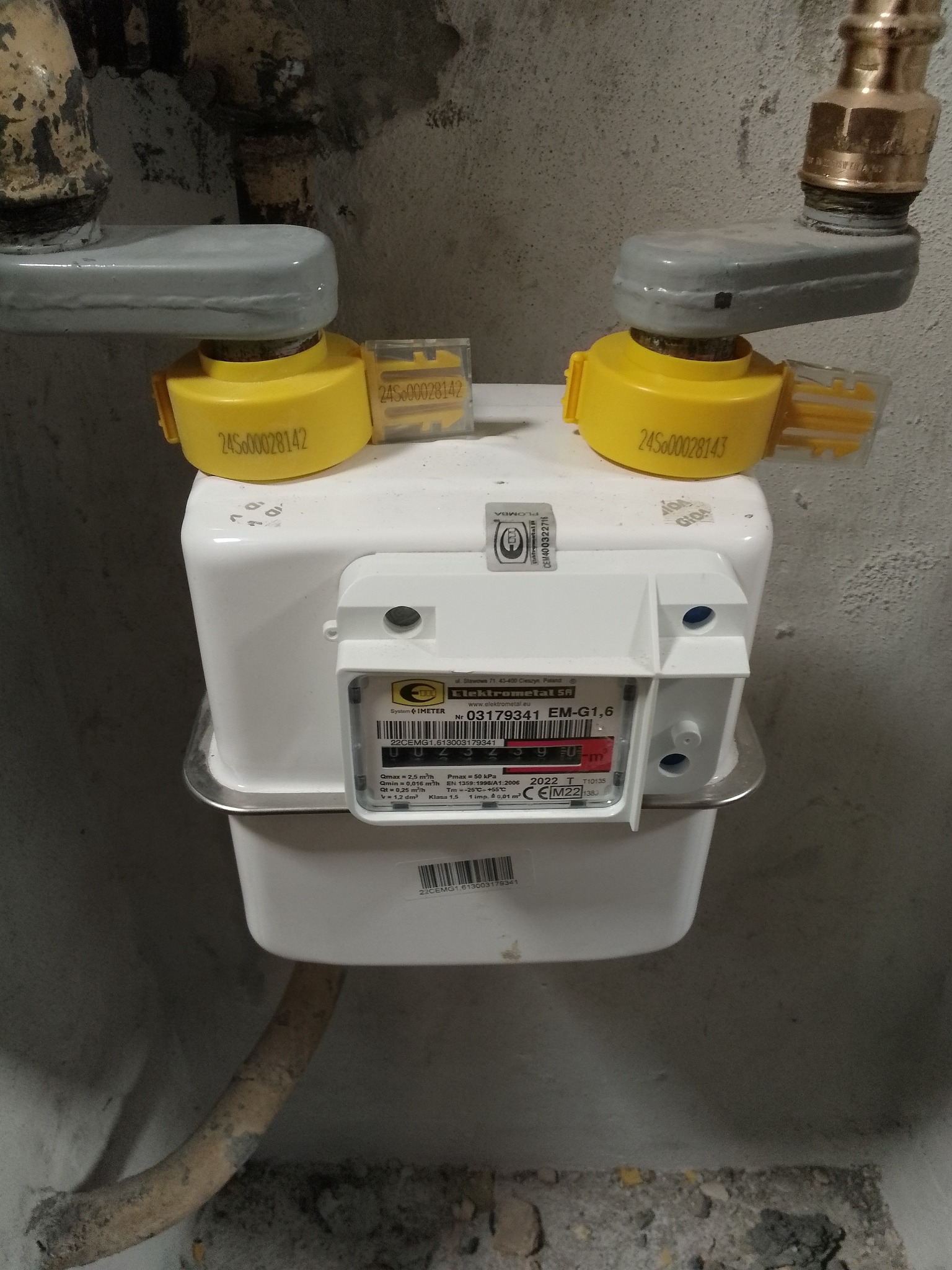 Zamknięty licznik gazu Elektrometal SA z żółtymi zaworami odcinającymi, zamontowany na szarej ścianie, widoczne rury doprowadzające gaz.