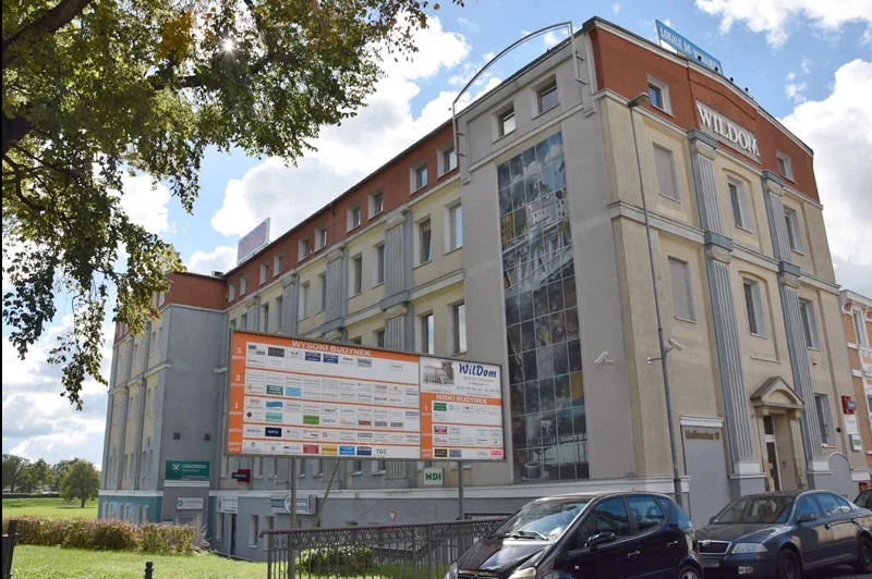 Budynek biurowy z reklamami firm, widoczny z poziomu ulicy, w tle błękitne niebo i drzewa. Na budynku szyld 'WILDOM' i duże okno z grafiką.