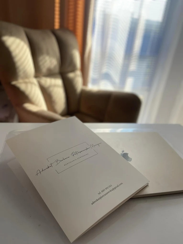 Kremowa teczka z danymi kontaktowymi adwokata, leżąca na biurku obok laptopa. W tle fotel i okno z jasnym światłem dziennym.