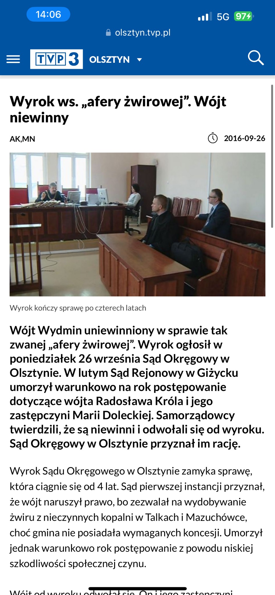 Wnętrze sali sądowej z sędziami za stołem i oskarżonymi na ławie, widoczne monitory komputerowe i drewniane elementy wyposażenia.