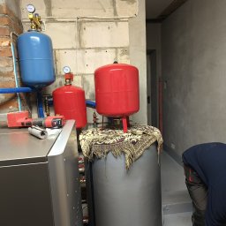 Piotr Orzechowicz - Instalacja centralnego ogrzewania w surowym wnętrzu: dwa czerwone zbiorniki wyrównawcze na wzorzystej tkaninie, niebieski zbiornik, nowoczesny piec, narzędzia i fragment sylwetki instalatora...