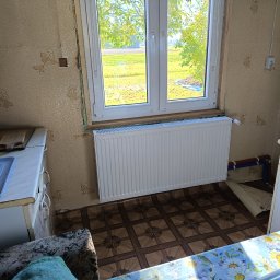 Piotr Orzechowicz - Biały grzejnik panelowy zamontowany pod oknem w pomieszczeniu z tapetą w kwiaty i brązową podłogą, widoczne rury instalacji grzewczej, okno z widokiem na zielone pole.