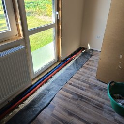 Piotr Orzechowicz - Instalacja grzewcza pod oknem: czerwona i niebieska rura prowadzone wzdłuż ściany, zabezpieczone folią, z widocznym grzejnikiem i widokiem na ogród za oknem.