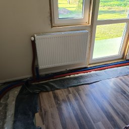 Piotr Orzechowicz - Biały grzejnik panelowy zamontowany przy oknie, z widocznymi czerwonymi i niebieskimi rurami instalacji centralnego ogrzewania, częściowo zakrytymi folią ochronną na drewnianej podłodze w trakcie...
