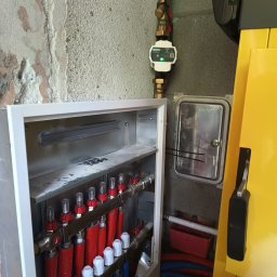 Piotr Orzechowicz - Rozdzielacz ogrzewania podłogowego w metalowej szafce, z widocznymi rurami w izolacji czerwonej i niebieskiej, zamontowany na surowej ścianie, obok żółtego kotła grzewczego.