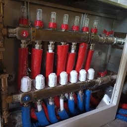 Piotr Orzechowicz - Rozdzielacz ogrzewania podłogowego w szafce, z widocznymi rurami w izolacji czerwonej i niebieskiej, przepływomierzami z czerwonymi pokrętłami, zaworami termostatycznymi z białymi głowicami...