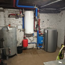 Piotr Orzechowicz - Piwnica z zainstalowanymi zbiornikami: ocynkowanym, białym i szarym, połączonymi rurami w izolacji czerwonej i niebieskiej, z czarnym wiadrem obok zbiornika ocynkowanego i pompą na podłodze.