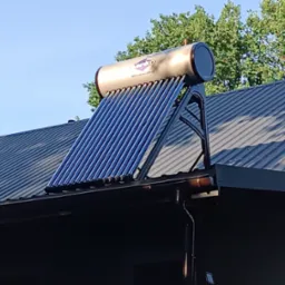 Kolektor słoneczny z rurami próżniowymi zamontowany na dachu z blachy trapezowej, widoczny z dołu pod kątem, w tle drzewa i błękitne niebo.