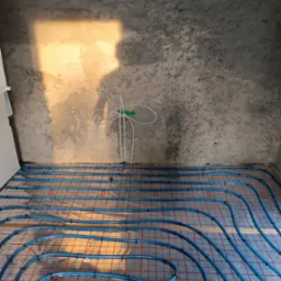 Niebieskie rury ogrzewania podłogowego ułożone na folii z czerwonym rastrem, widoczne białe rurki i cień okna na surowej ścianie w tle.