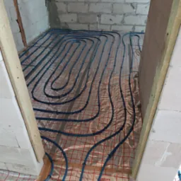 Układanie niebieskich rur ogrzewania podłogowego na folii izolacyjnej z siatką w surowym pomieszczeniu z bloczków betonowych, widok z góry przez futrynę.