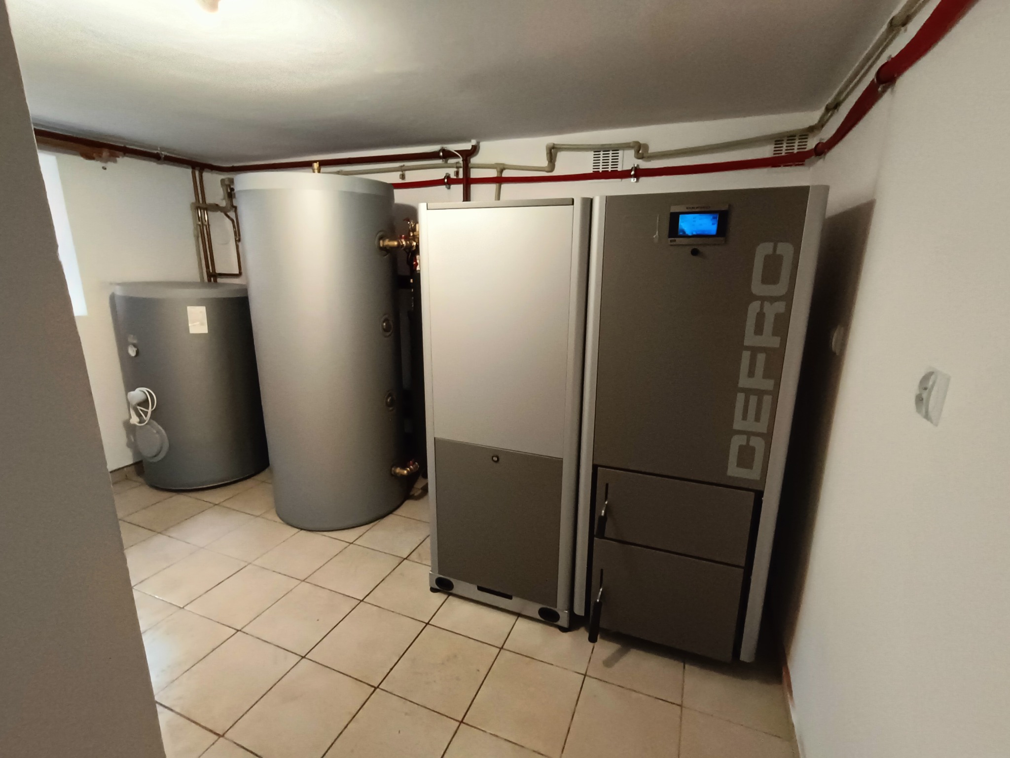Kotłownia z dwoma zbiornikami akumulacyjnymi i piecem DEFRO, widoczne rury instalacji grzewczej pod sufitem, płytki podłogowe w beżowym odcieniu.