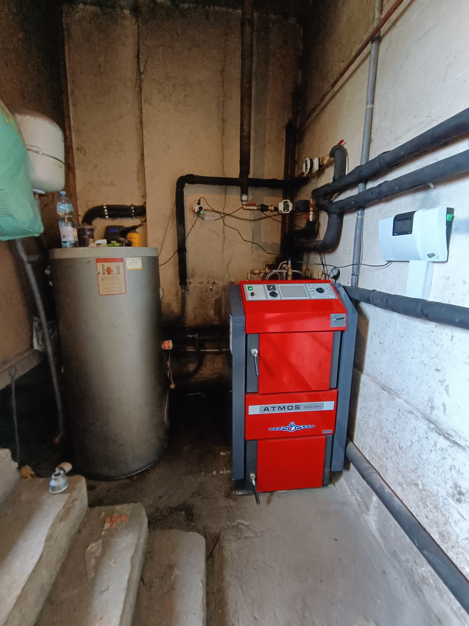 Czerwono-szary piec grzewczy Atmos Generator w kotłowni, obok srebrny bojler, widoczne rury instalacji, schody betonowe w tle brudne ściany.