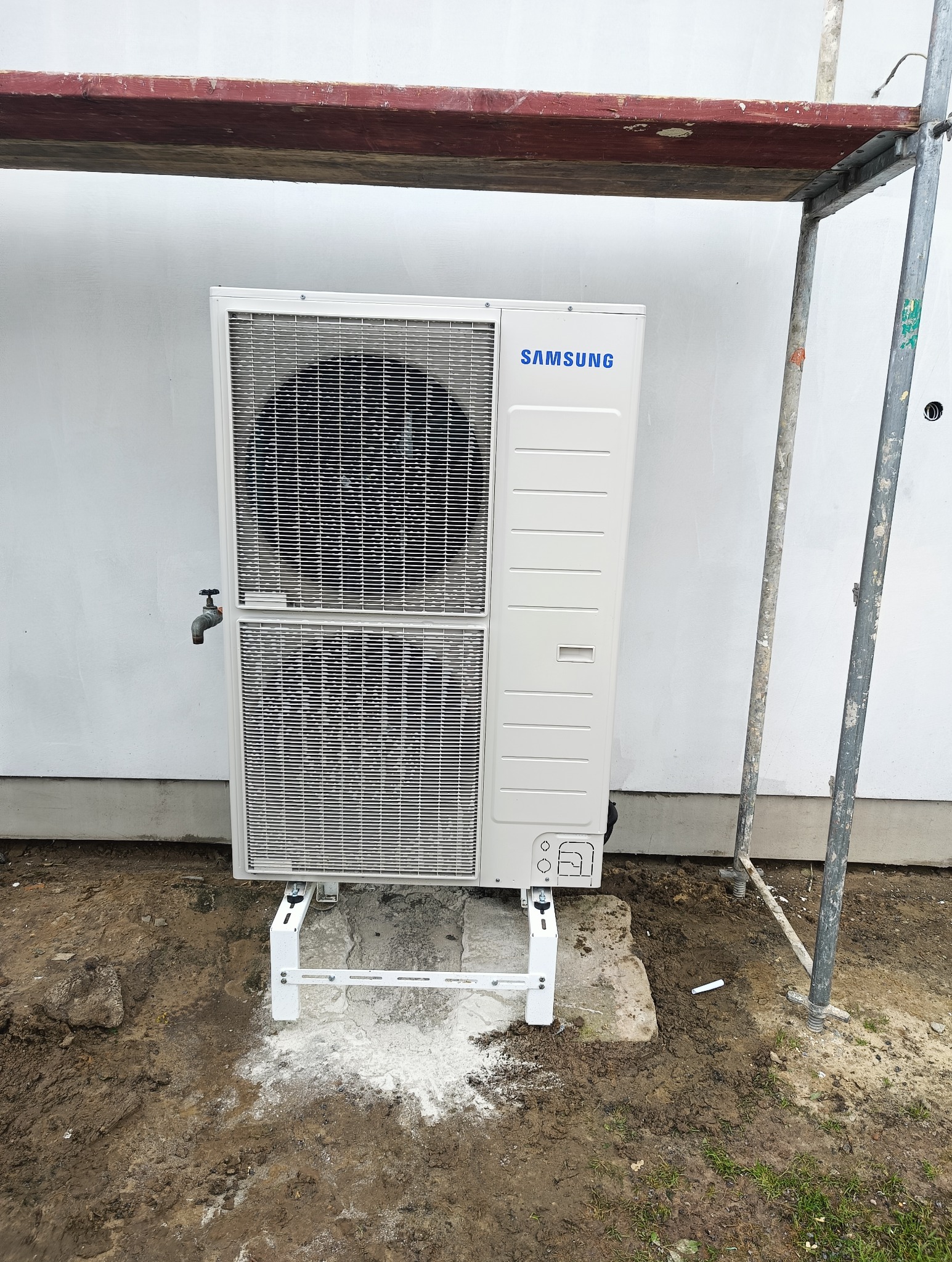 Biała zewnętrzna jednostka pompy ciepła Samsung zamontowana na betonowym fundamencie, widoczne rusztowanie i kran na ścianie budynku.