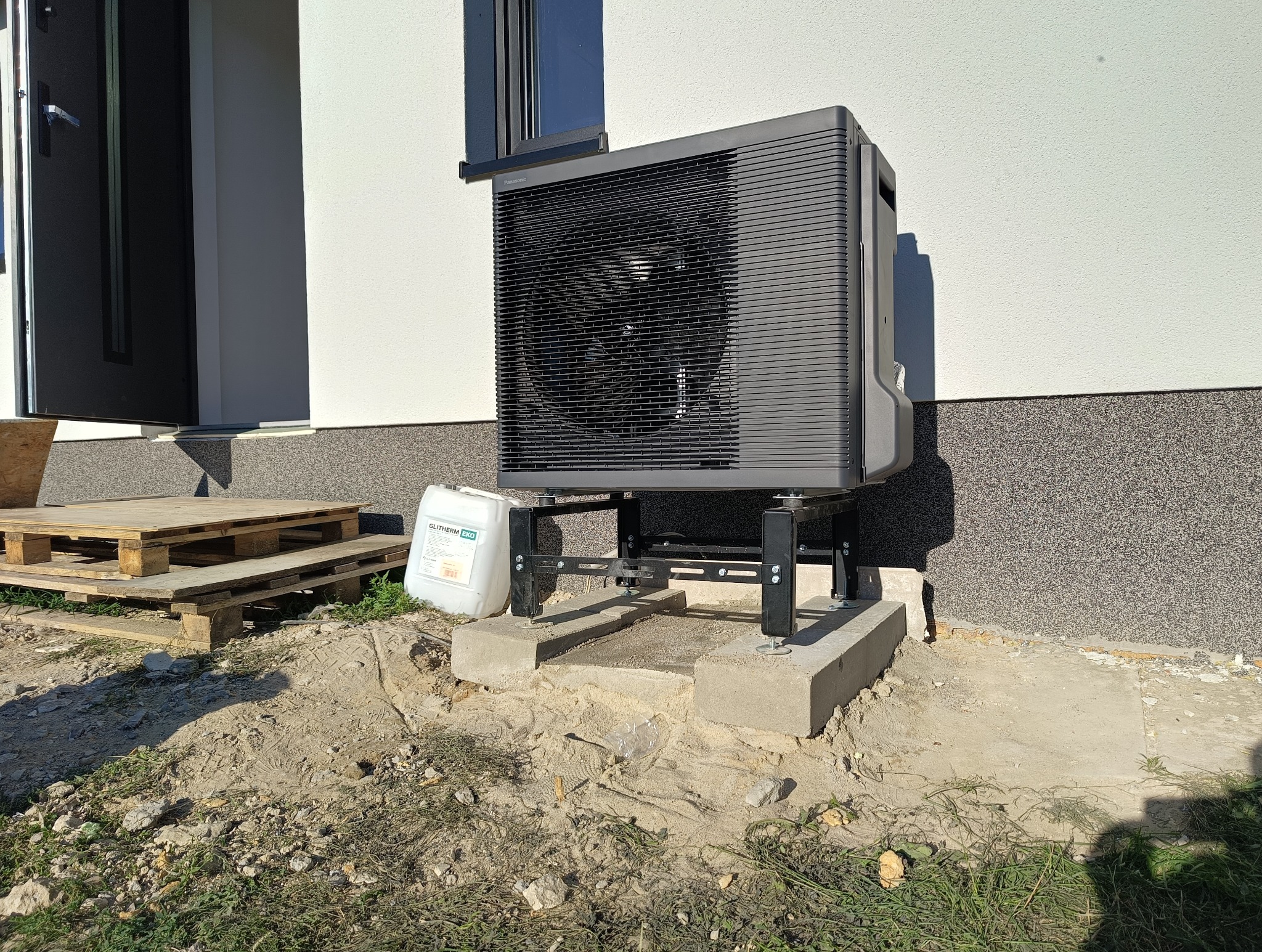 Zewnętrzna jednostka pompy ciepła Panasonic Aquarea zamontowana na betonowym fundamencie, obok paleta drewniana i kanister z płynem Glitherm EKO.
