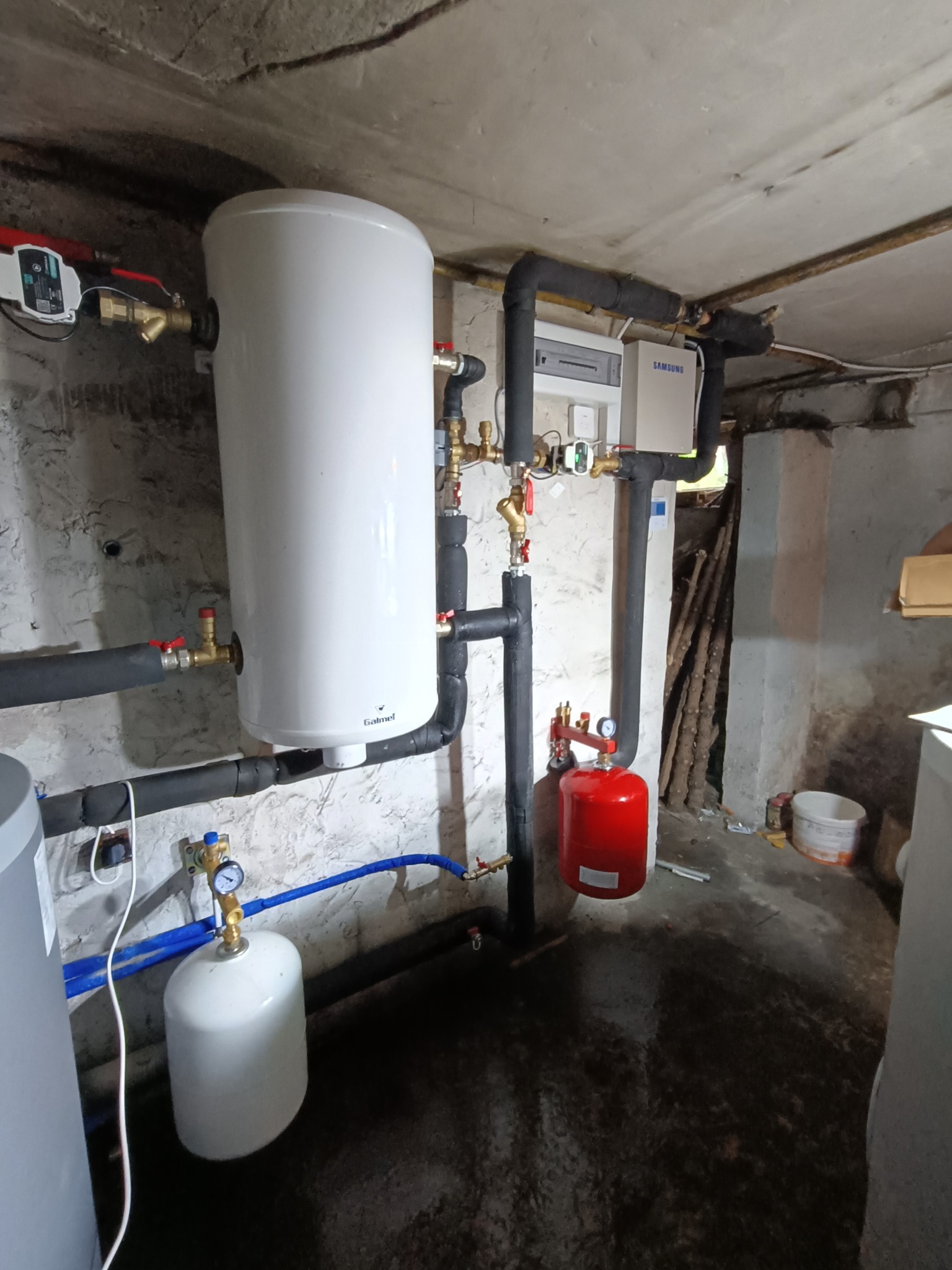 Biały zasobnik ciepłej wody Galmet z czarnymi rurami izolowanymi termicznie, czerwony zbiornik przeponowy i biała skrzynka rozdzielcza Samsung na tle surowej ściany piwnicy.