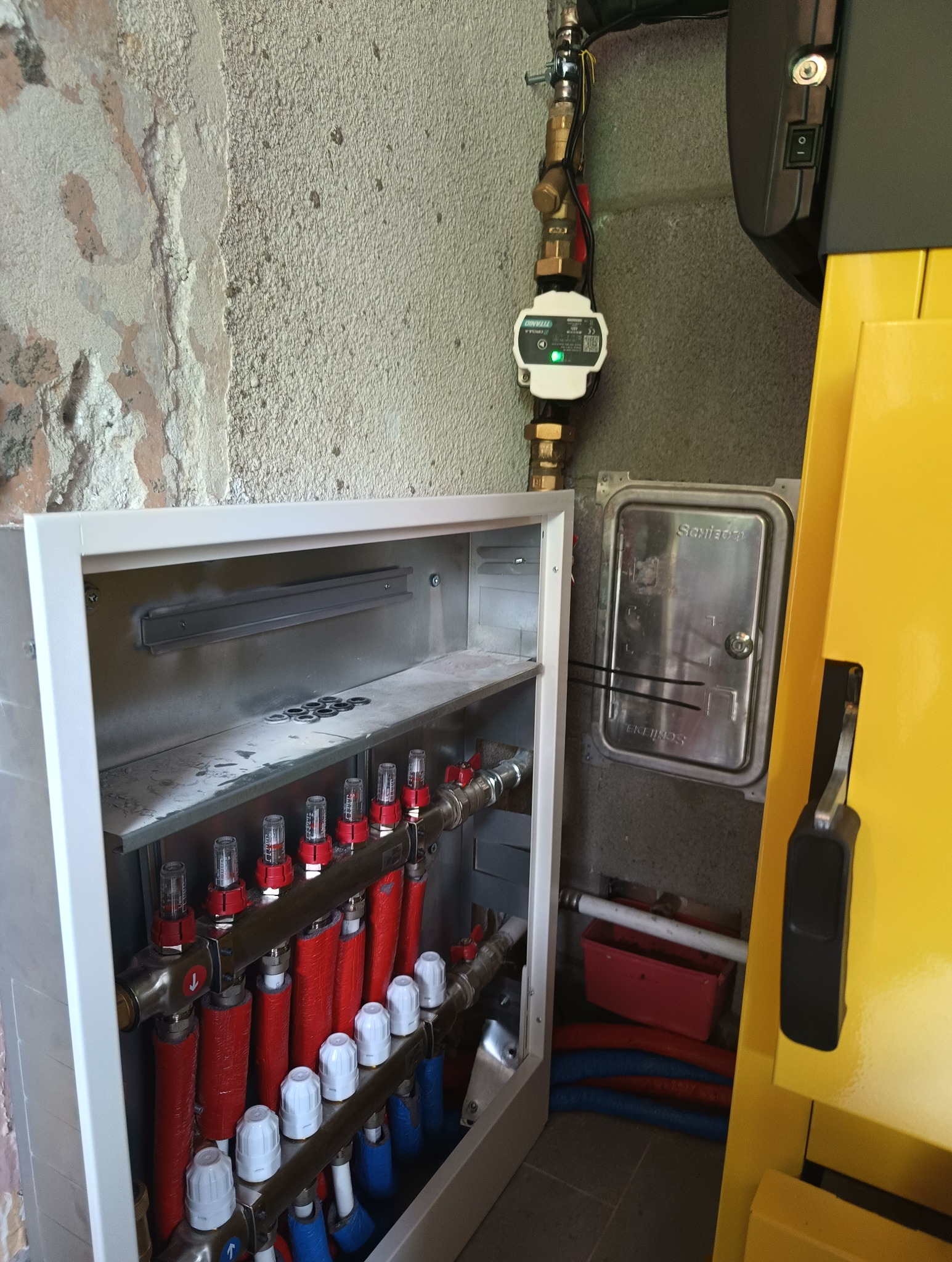 Rozdzielacz ogrzewania podłogowego w metalowej szafce, z widocznymi rurami w izolacji czerwonej i niebieskiej, zamontowany na surowej ścianie, obok żółtego kotła grzewczego.