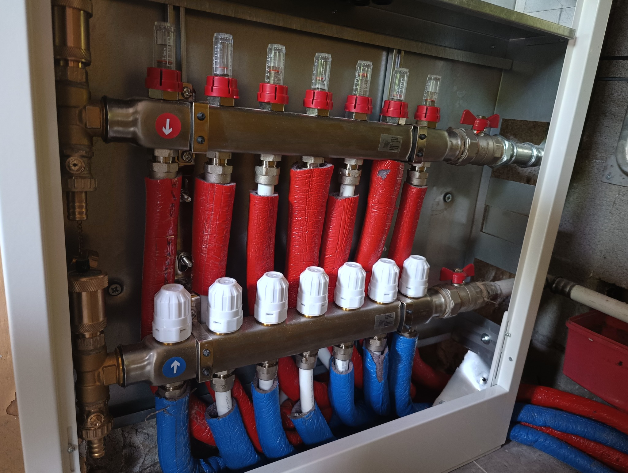 Rozdzielacz ogrzewania podłogowego w szafce, z widocznymi rurami w izolacji czerwonej i niebieskiej, przepływomierzami z czerwonymi pokrętłami, zaworami termostatycznymi z białymi głowicami...