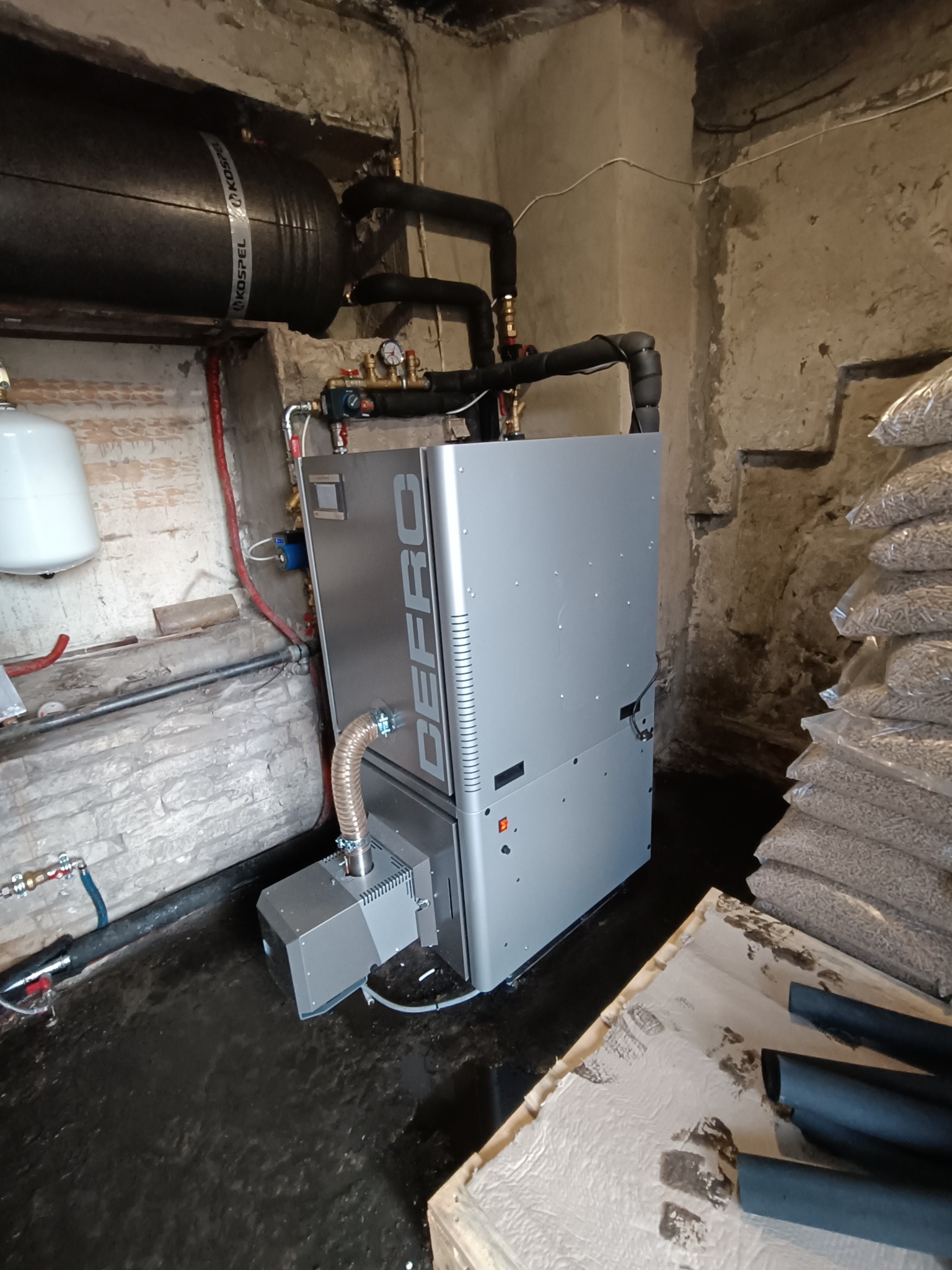 Nowoczesny piec na pellet marki DEFRO w kotłowni, z widocznym zasobnikiem Kospel, workami pelletu obok i instalacją hydrauliczną, w surowym wnętrzu.
