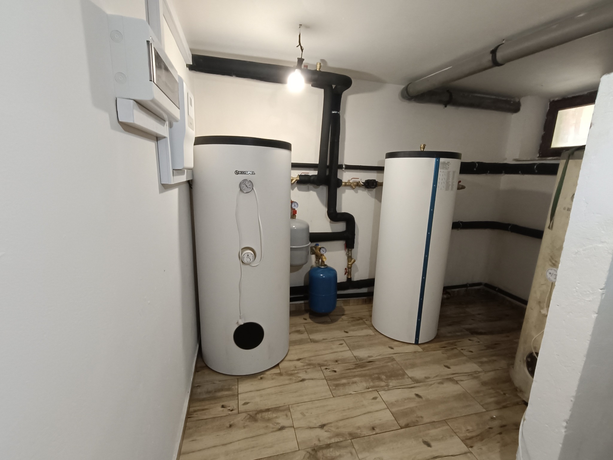 Dwa białe zbiorniki akumulacyjne ciepłej wody w kotłowni, widoczne rury grzewcze w izolacji, niebieski zbiornik wyrównawczy i ceramiczna podłoga imitująca drewno.