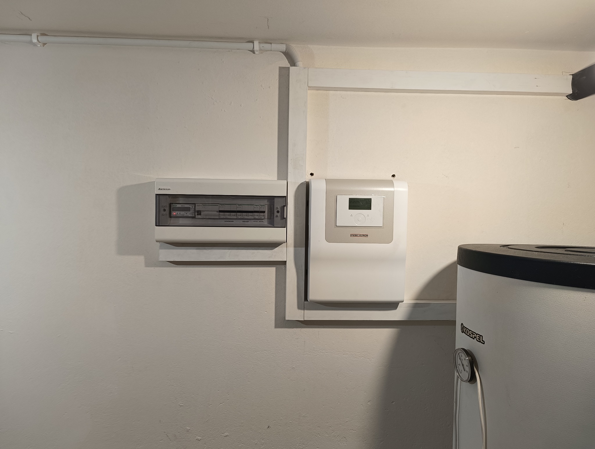 Biała ściana z zamontowaną skrzynką bezpiecznikową, regulatorem Stiebel Eltron i fragmentem bojlera Kospel z termometrem analogowym. Widoczne rurki instalacyjne biegnące wzdłuż ściany.