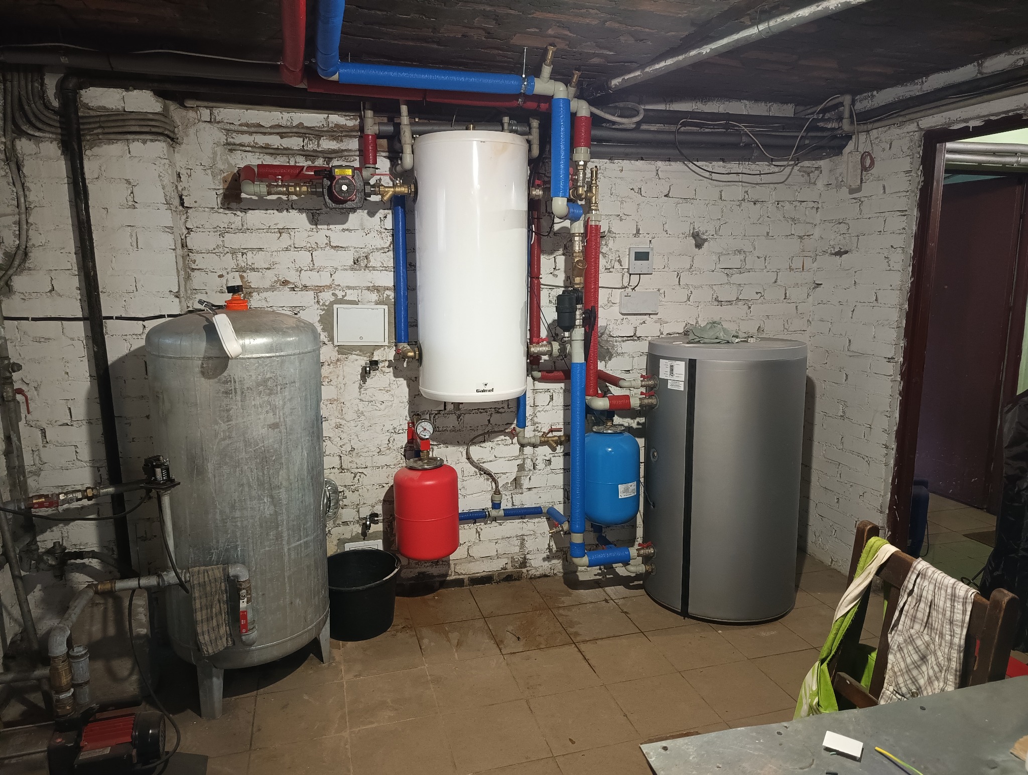 Piwnica z zainstalowanymi zbiornikami: ocynkowanym, białym i szarym, połączonymi rurami w izolacji czerwonej i niebieskiej, z czarnym wiadrem obok zbiornika ocynkowanego i pompą na podłodze.
