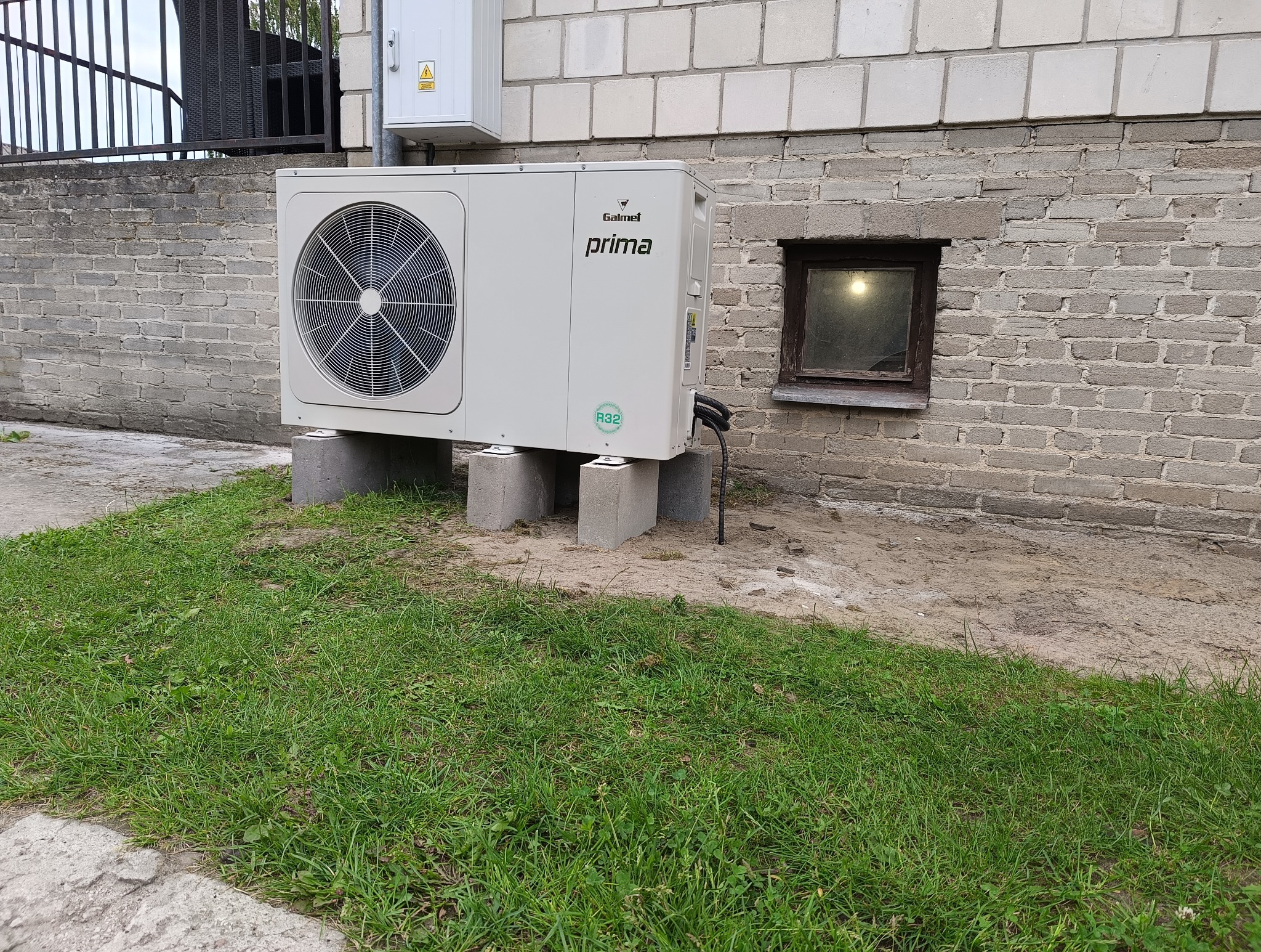 Zewnętrzna jednostka klimatyzacji Galmet Prima R32 umieszczona na betonowych bloczkach przy ceglanej ścianie z małym okienkiem piwnicznym.