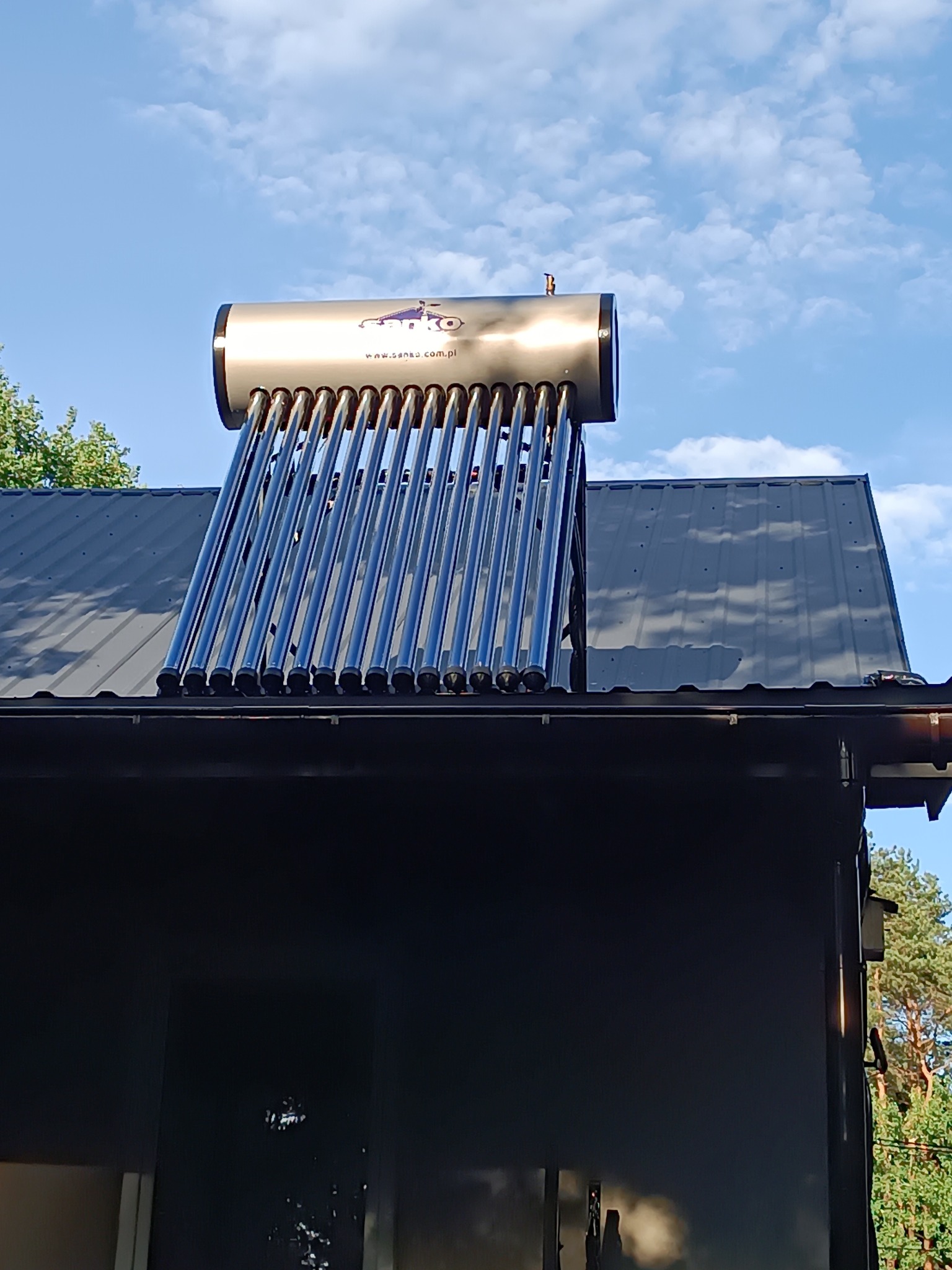 Kolektor słoneczny rurowo-próżniowy zamontowany na dachu budynku, widoczny zbiornik ze stali nierdzewnej z logo producenta, błękitne niebo w tle.