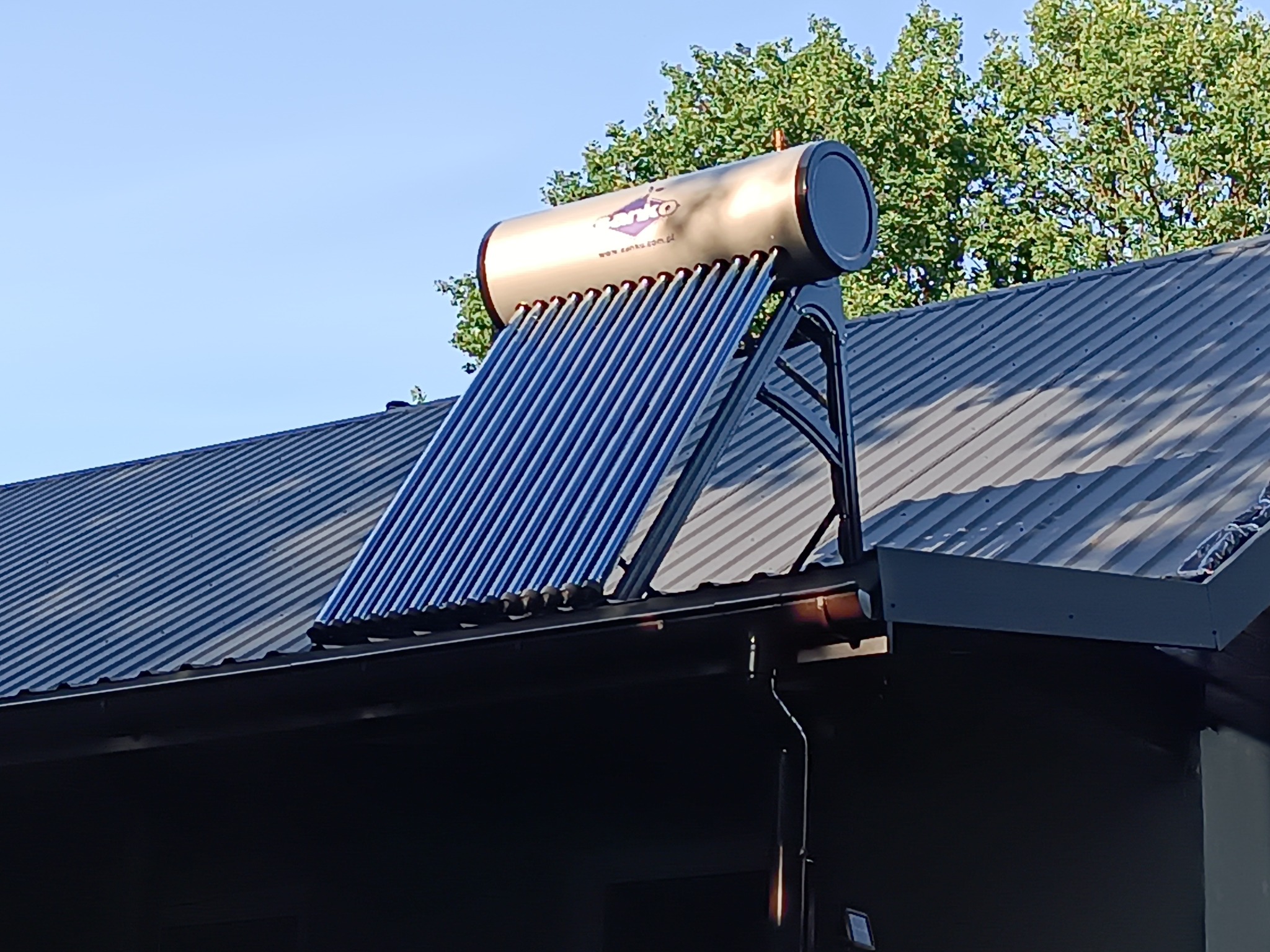 Kolektor słoneczny z rurami próżniowymi zamontowany na dachu z blachy trapezowej, widoczny z dołu pod kątem, w tle drzewa i błękitne niebo.