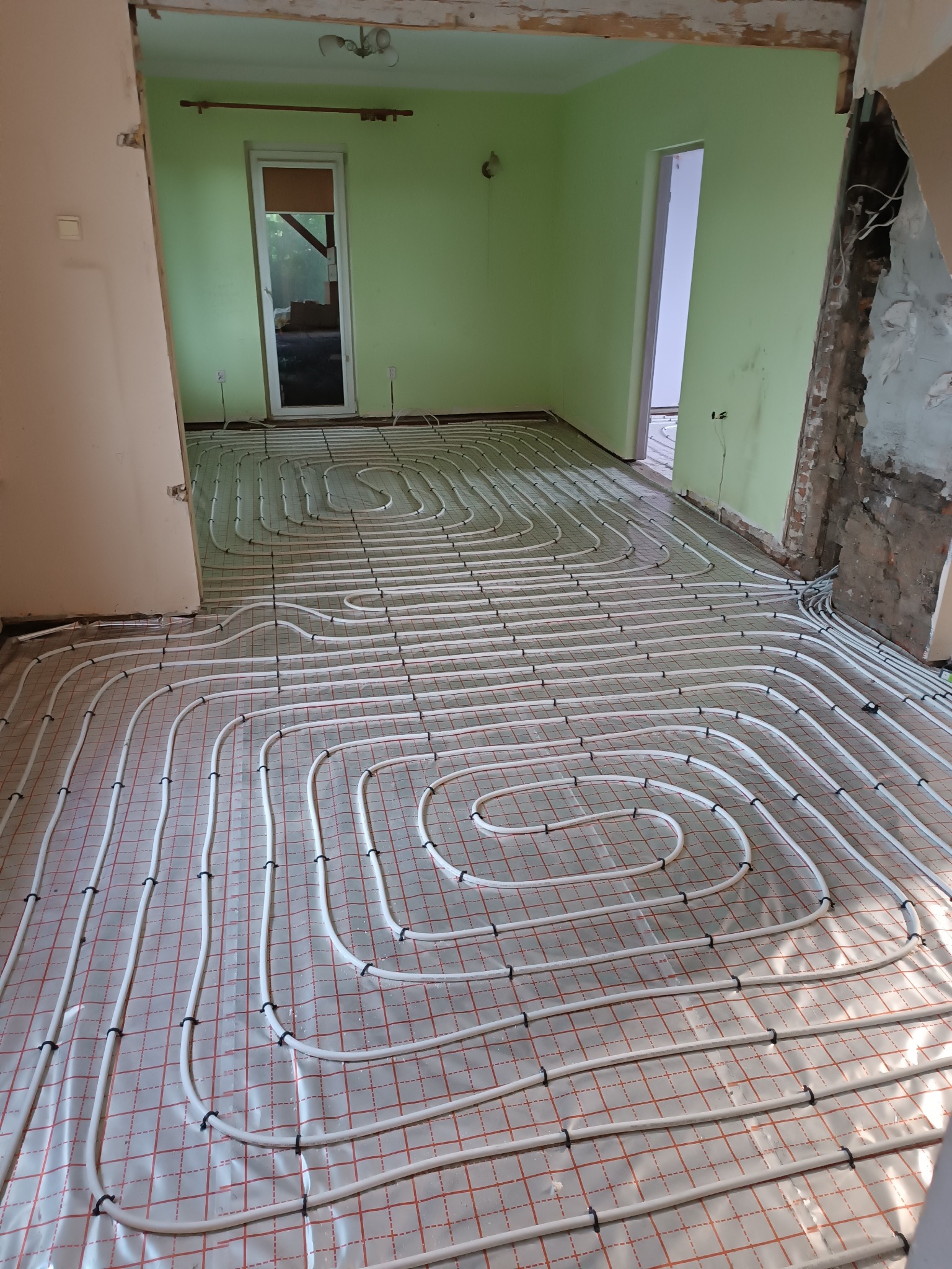 Białe rury ogrzewania podłogowego zamocowane na folii z czerwoną siatką, widok na surowy stan ścian i zielone ściany w tle.