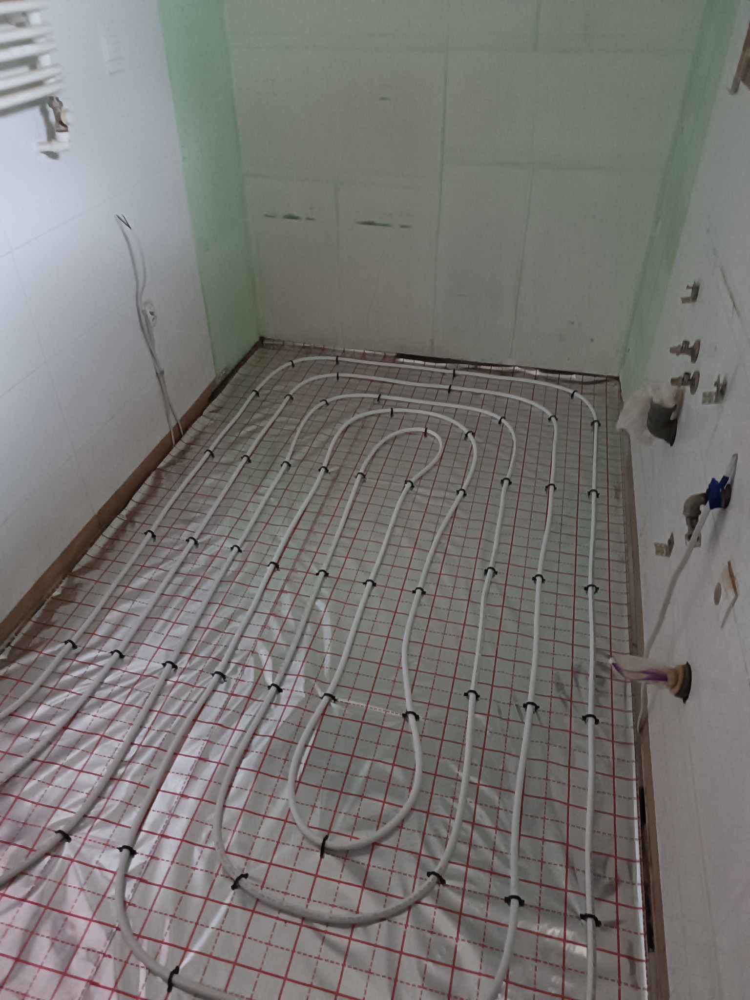 Białe rury ogrzewania podłogowego ułożone na folii z czerwoną kratką w surowym wnętrzu łazienki z zielonymi akcentami, widoczne przyłącza na ścianie.