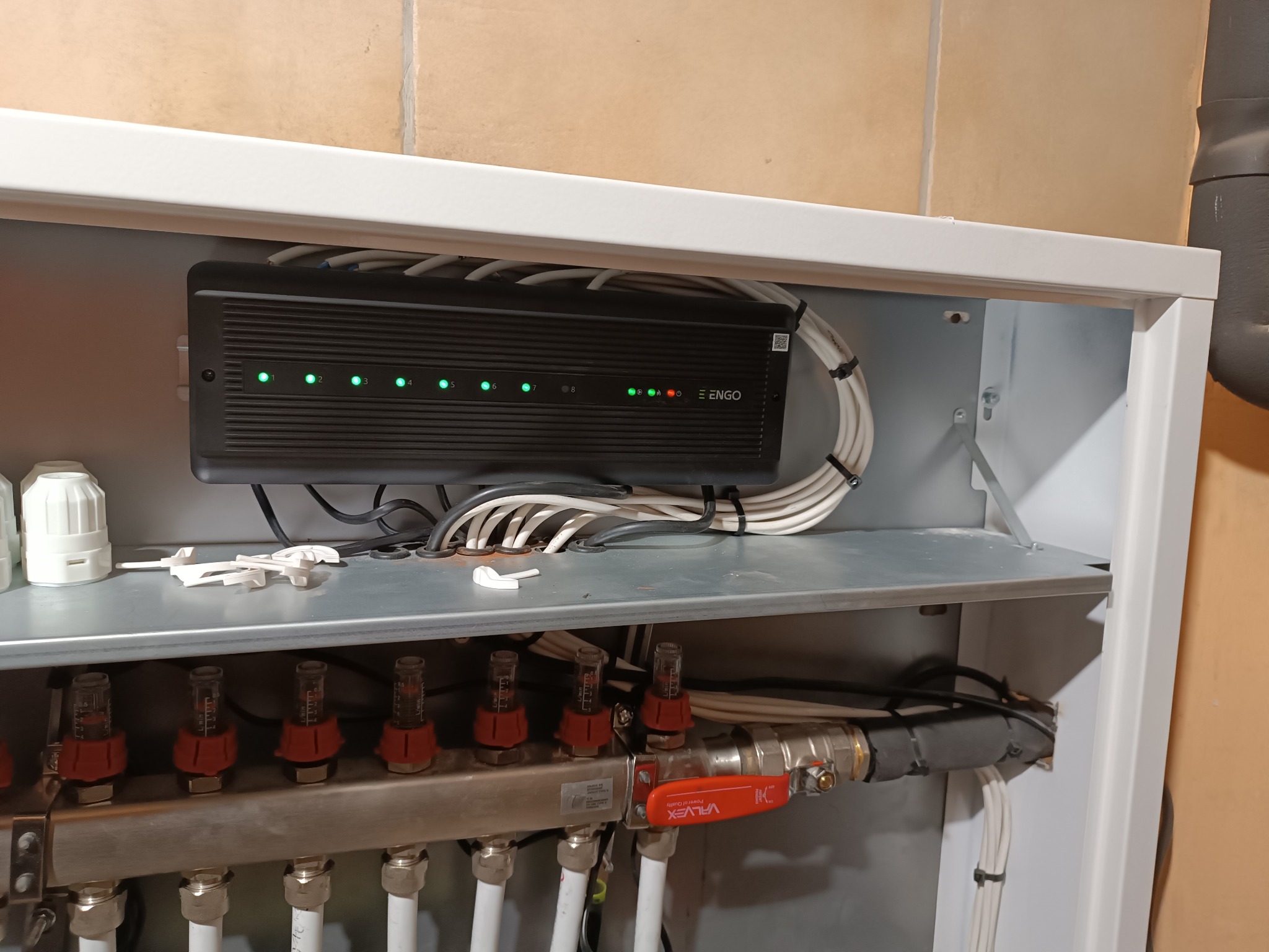 Rozdzielacz ogrzewania podłogowego w szafce, widoczne rurki, zawory, przepływomierze z czerwonymi elementami, regulator ENGO z diodami LED, białe kable spięte opaskami.