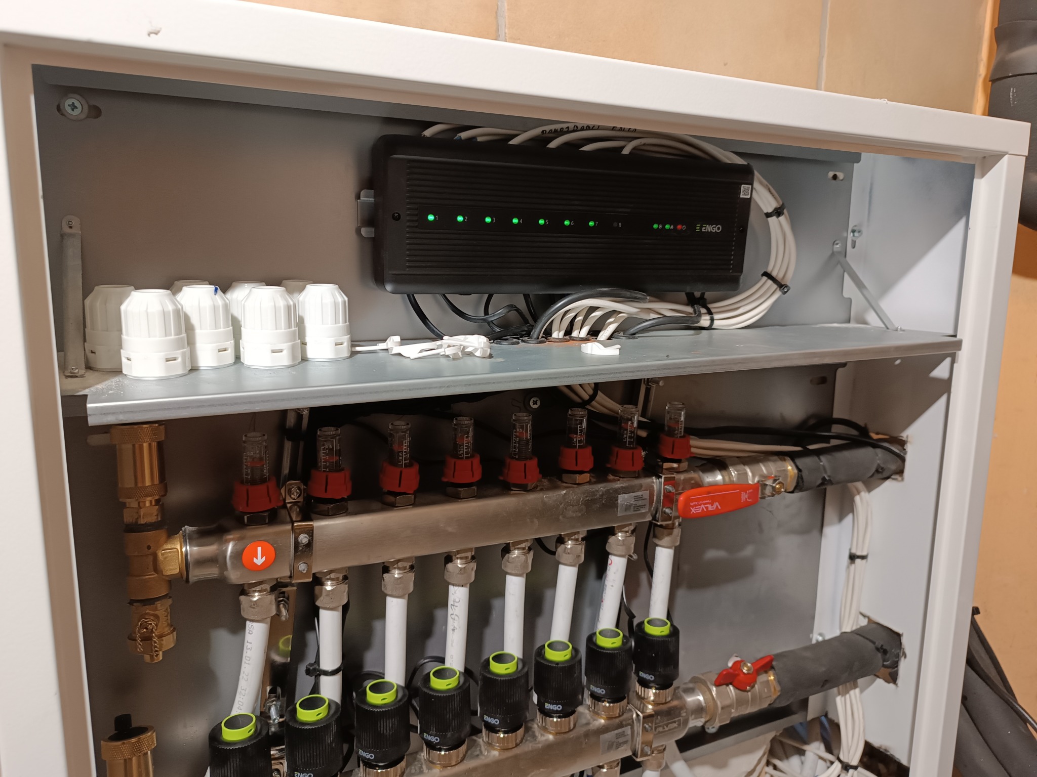 Rozdzielacz podłogowy ENGO w szafce z widocznym sterownikiem i przewodami, z zielonymi diodami sygnalizacyjnymi, rurami i zaworami, zainstalowany przy beżowej ścianie.