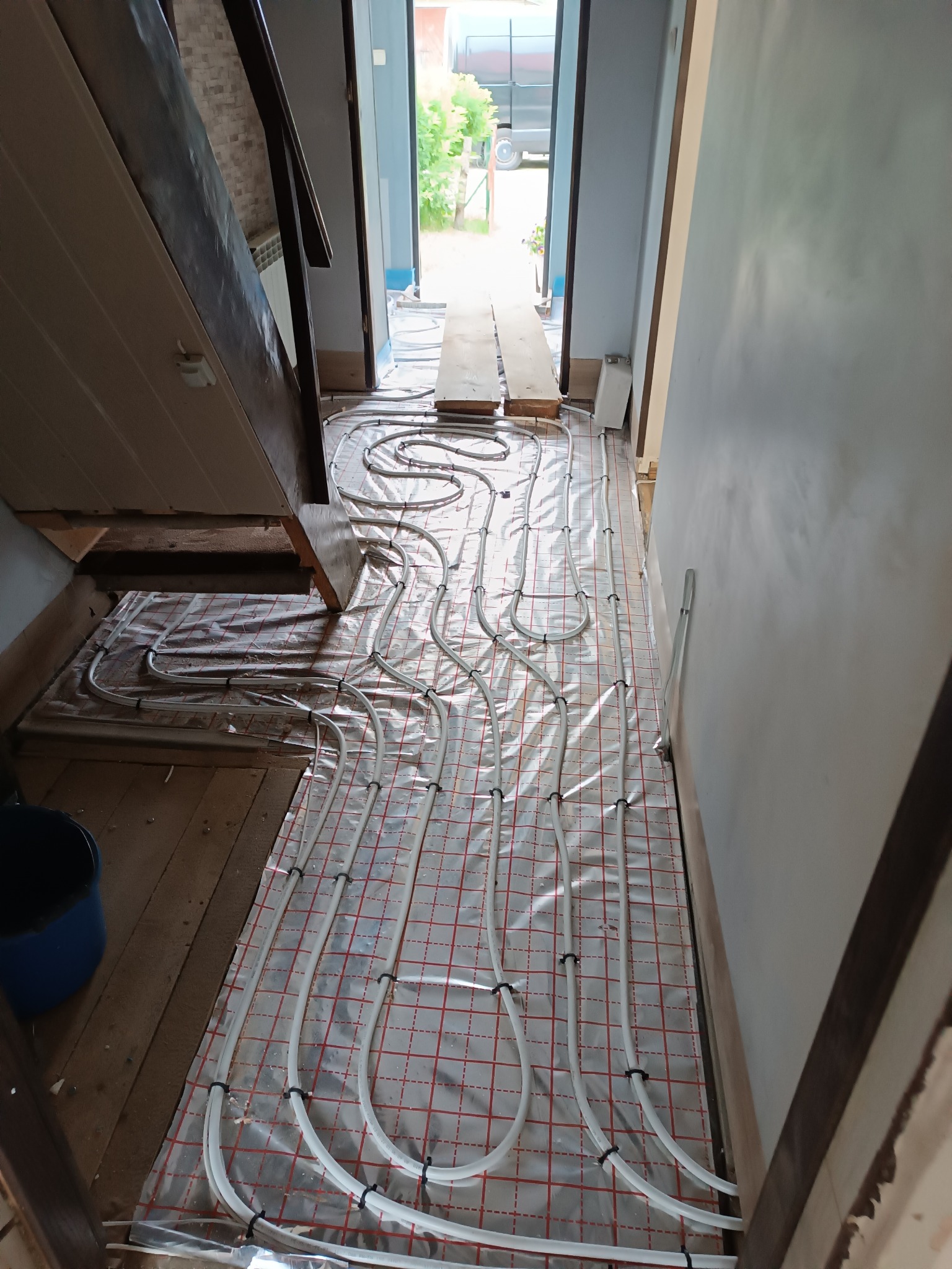 Białe rury ogrzewania podłogowego, przymocowane czarnymi opaskami do folii z czerwoną kratką, rozłożone na drewnianej podłodze w trakcie montażu, widok z góry, w tle schody i otwarte drzwi.