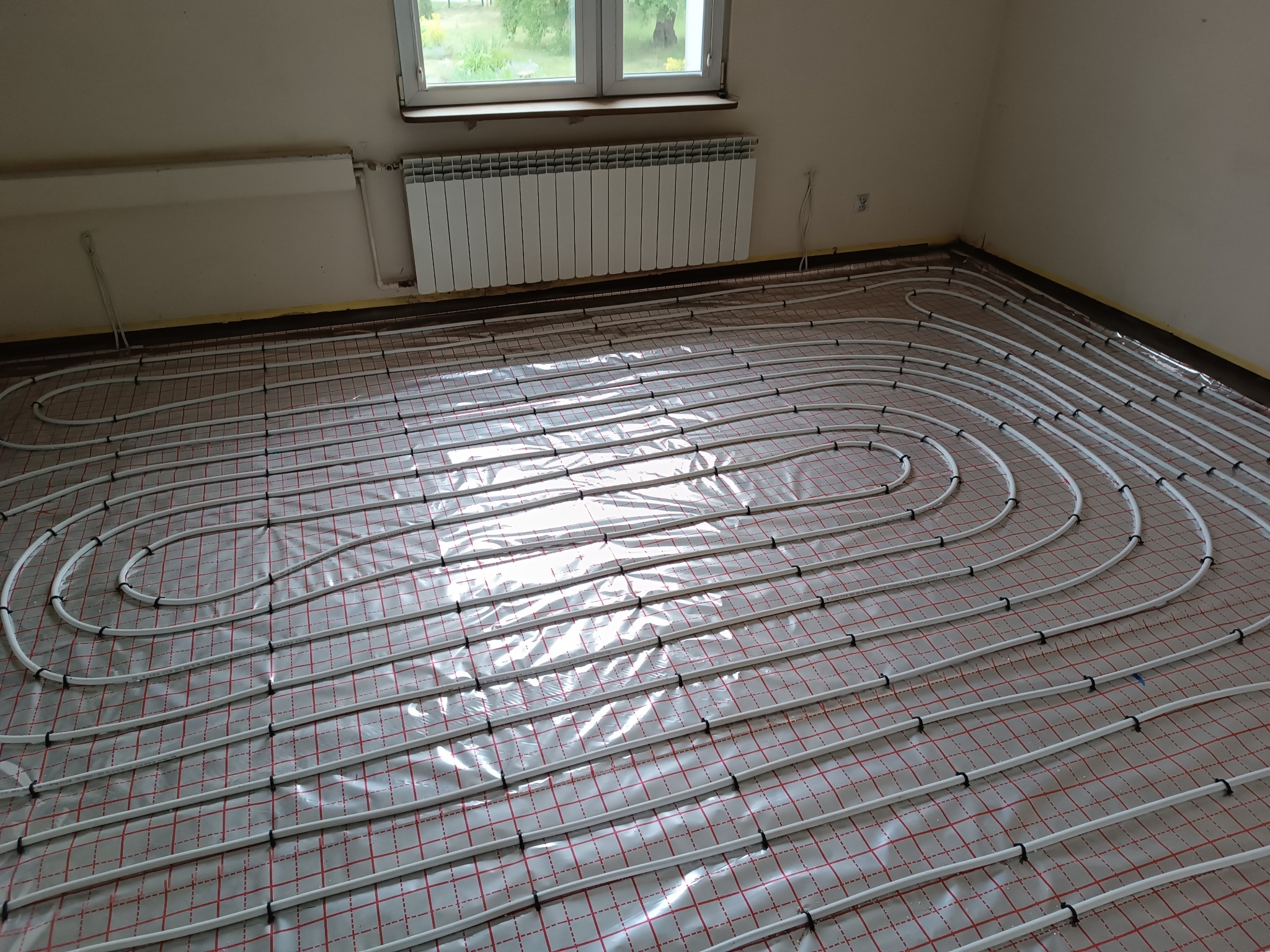 Białe rurki ogrzewania podłogowego ułożone spiralnie na folii z czerwoną siatką w jasnym pomieszczeniu, widoczny grzejnik ścienny.