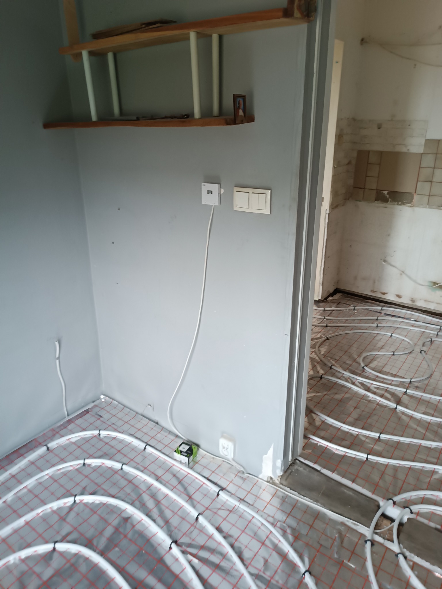 Białe rury ogrzewania podłogowego rozłożone na czerwonej siatce w szarym pomieszczeniu w trakcie instalacji, widoczny termostat na ścianie i półki w tle