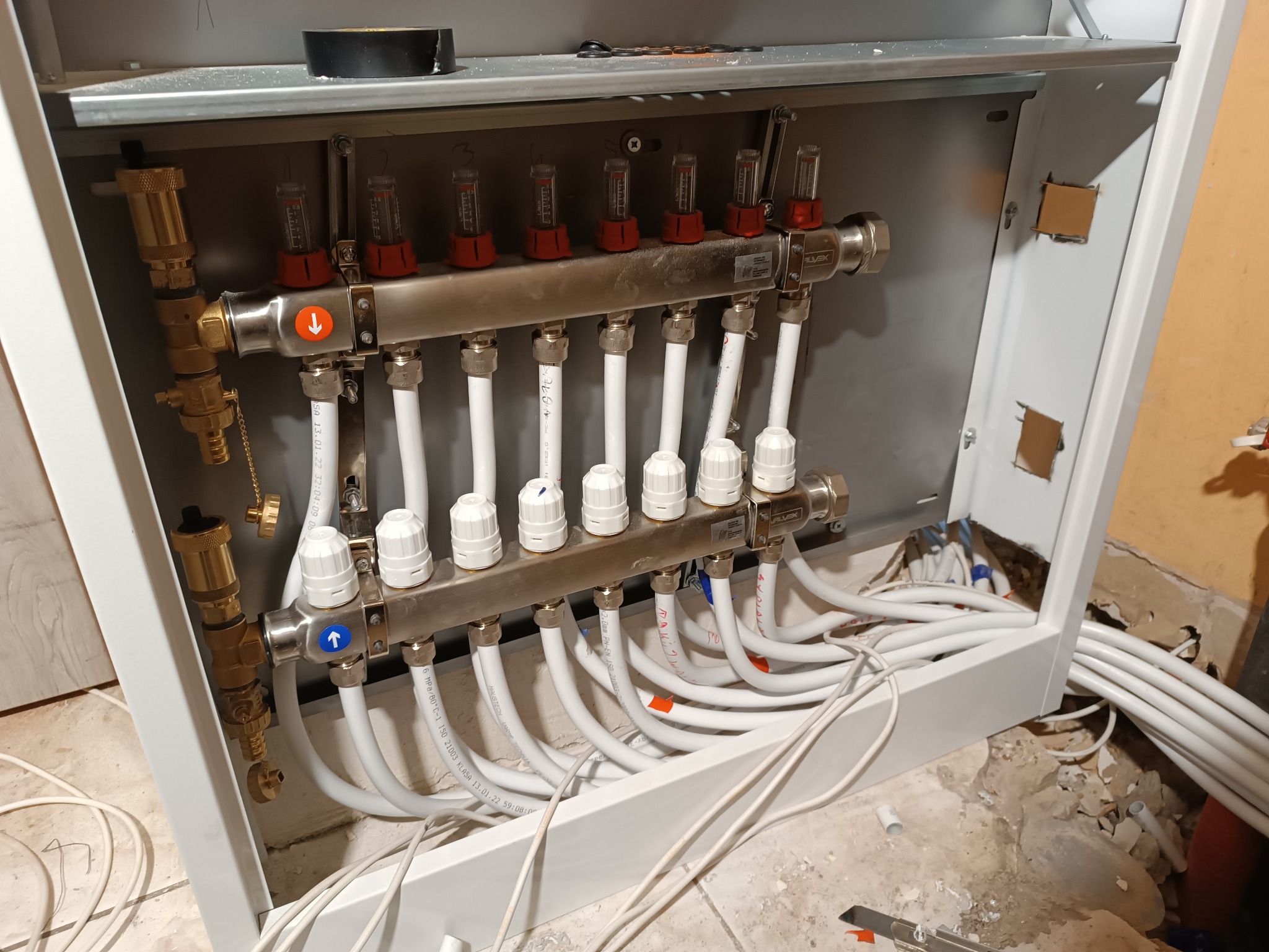 Rozdzielacz ogrzewania podłogowego w metalowej szafce, widoczne zawory, rury i przepływomierze, instalacja w trakcie montażu, widoczne przewody i fragment niedokończonej ściany.