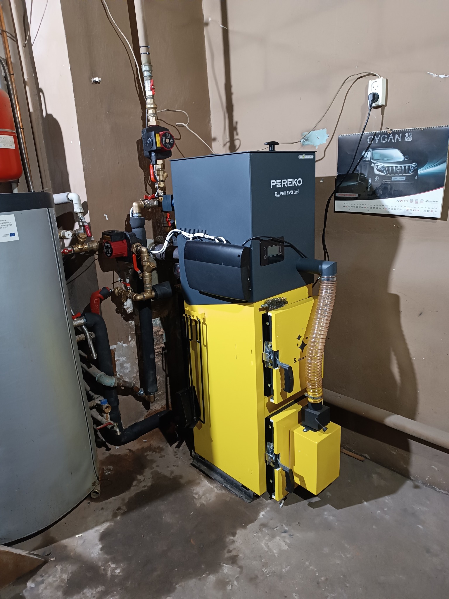 Żółto-szary piec na pellet marki PEREKO QmPell EVO BR z podłączonymi rurami, widoczny kalendarz z Toyotą Land Cruiser na ścianie, pomieszczenie z widocznymi śladami wilgoci na podłodze.