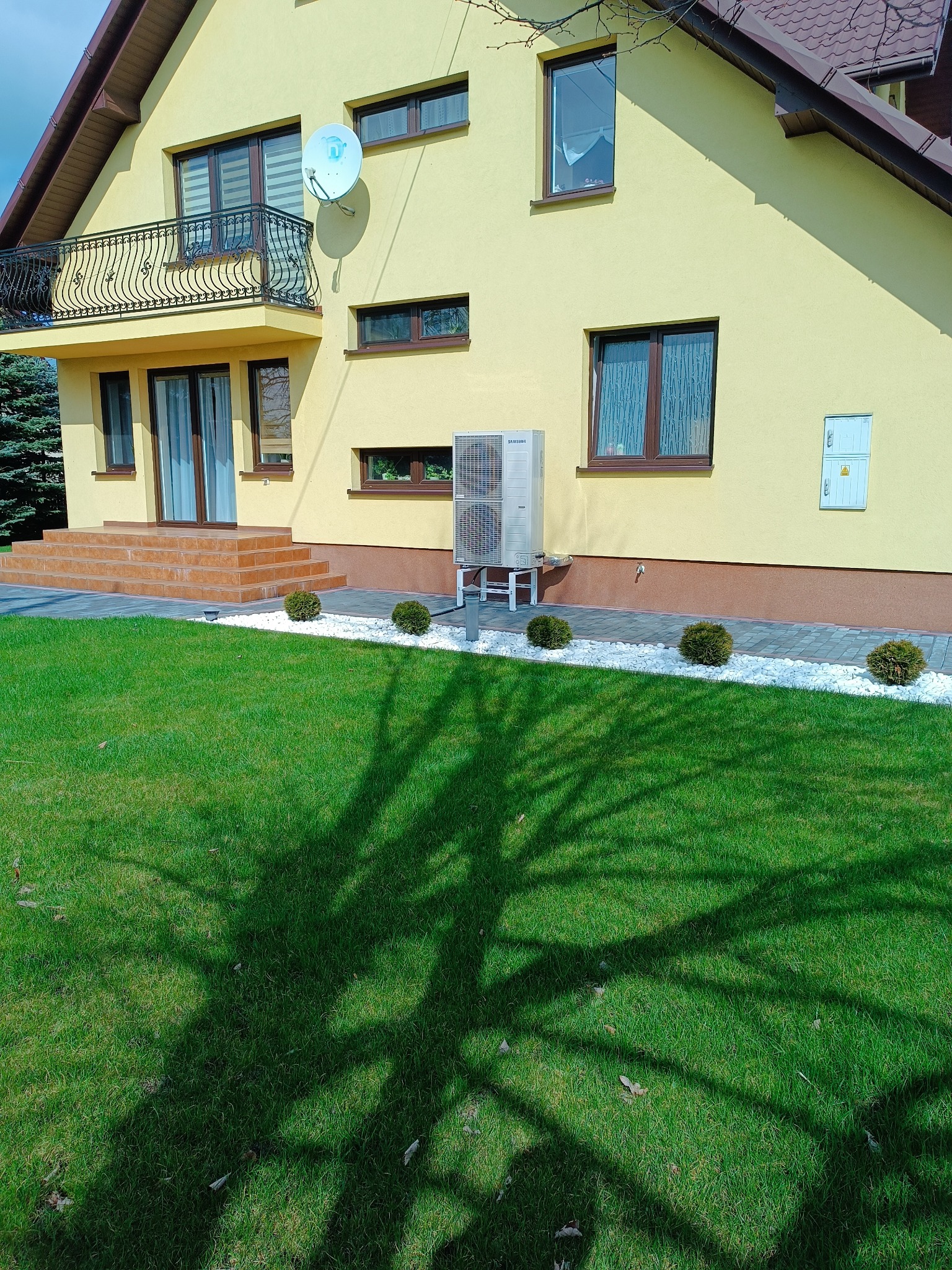 Żółty dom jednorodzinny z zamontowaną jednostką zewnętrzną klimatyzacji Samsung na białym podeście, widok z trawnika z cieniem drzewa.