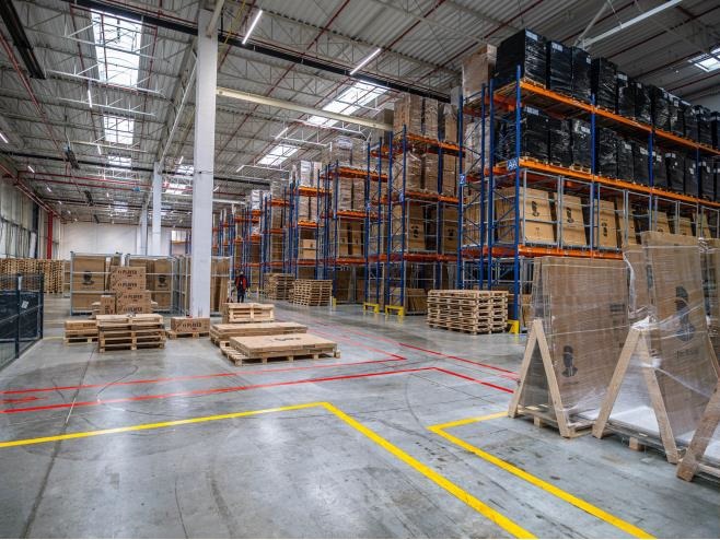Wnętrze magazynu z wysokimi regałami wypełnionymi kartonami i czarnymi opakowaniami, paletami na podłodze z namalowanymi żółtymi i czerwonymi liniami, oraz pracownikiem w oddali.