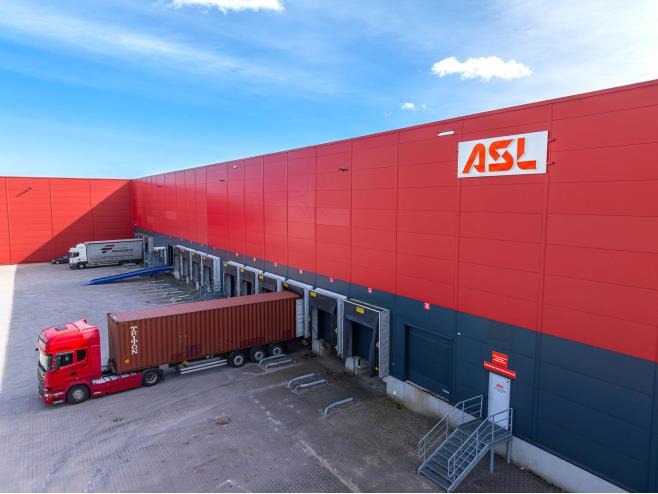 Czerwona ciężarówka z kontenerem Triton przy rampie załadunkowej magazynu ASL z czerwono-szarymi ścianami, widok z góry pod lekkim kątem.