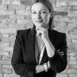 Czarno-białe portretowe ujęcie kobiety w czarnej marynarce, z ręką pod brodą i skrzyżowanymi ramionami, na tle ściany z cegieł